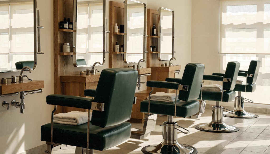 Four green barber chairs beside mirrors and oak workstations in a sunlit shop.